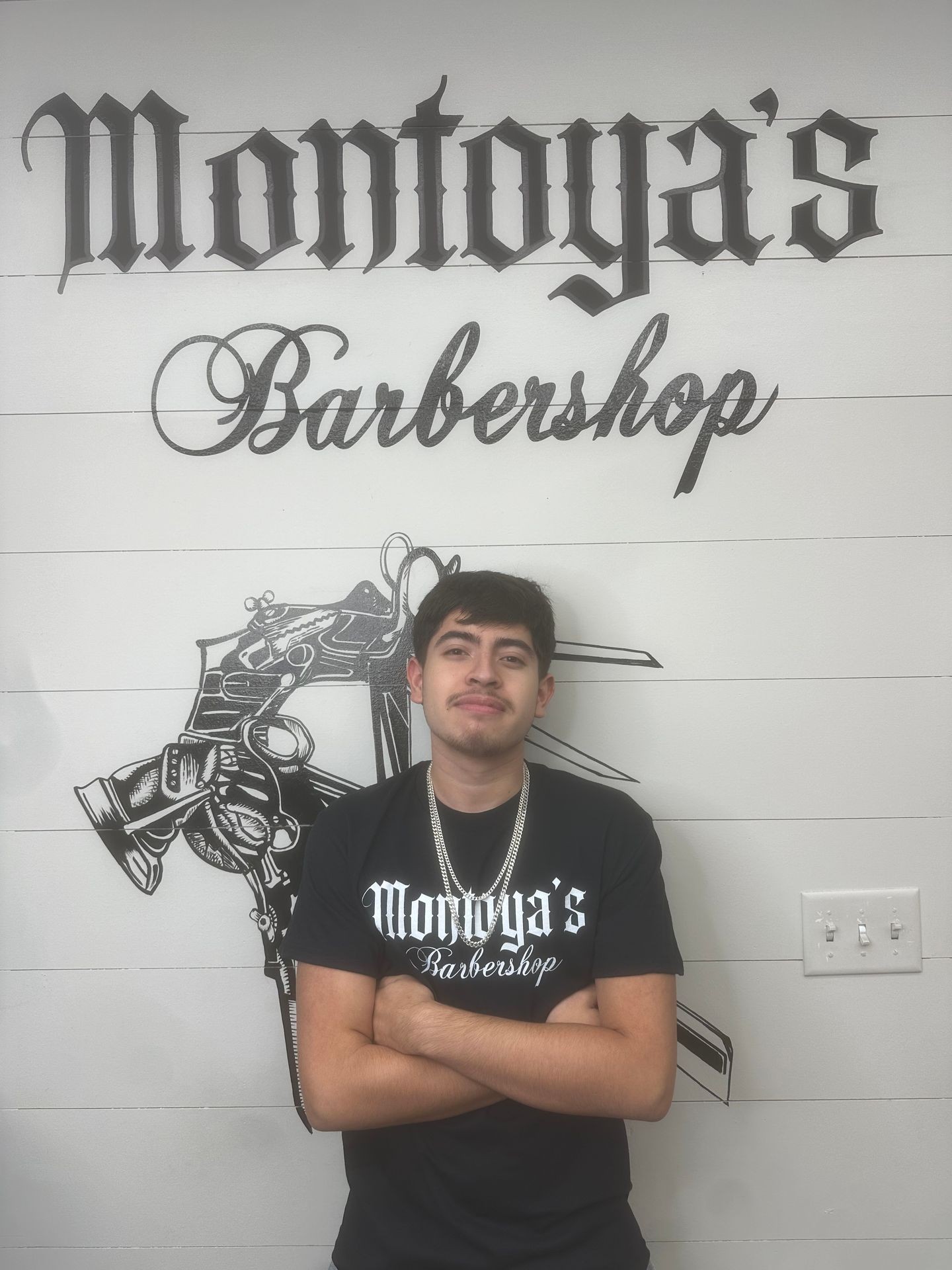 Person standing in front of Montoya's Barbershop sign, wearing a branded t-shirt.