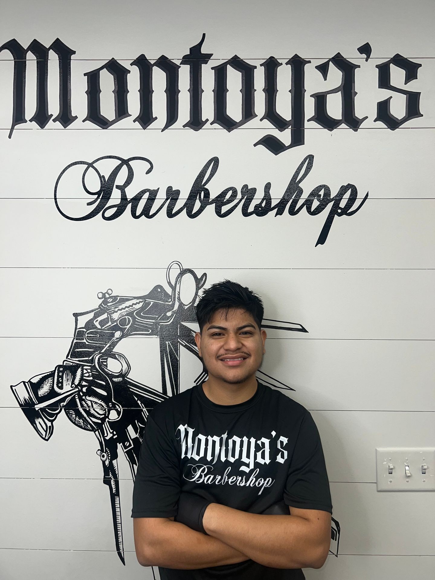 Person smiling in front of Montoya's Barbershop logo on a wall.